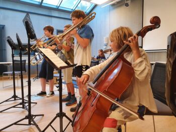 5 Kinder machen gemeinsam Musik mit einem Cello, einer Posaune, zwei Trompeten und einem Schlagzeug.