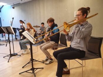 Im Nordkolleg sitzen 5 Jugendliche mit verschiedenen Instrumenten (Posaune, Saxophon, Drumset, E-Bass und Gesang) nebeneinander und musizieren zusammen.